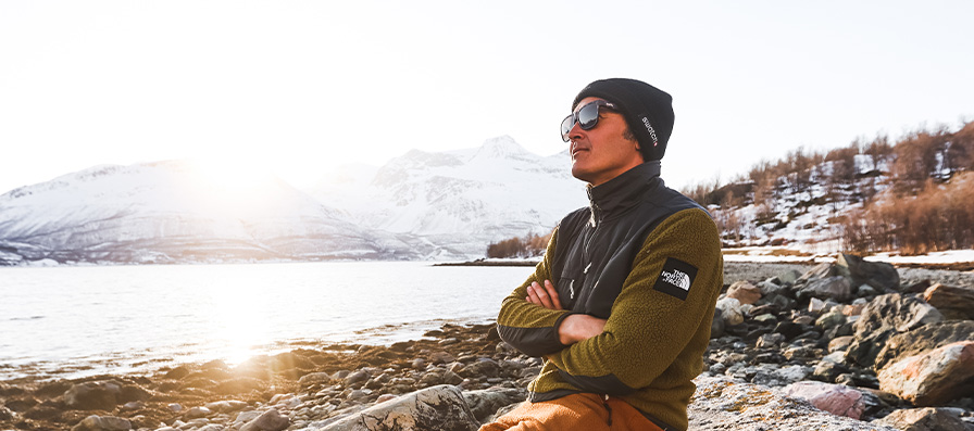 Man sitting on a rock by a lake in winter wearing Lowdown 2 Sunglasses and a beanie